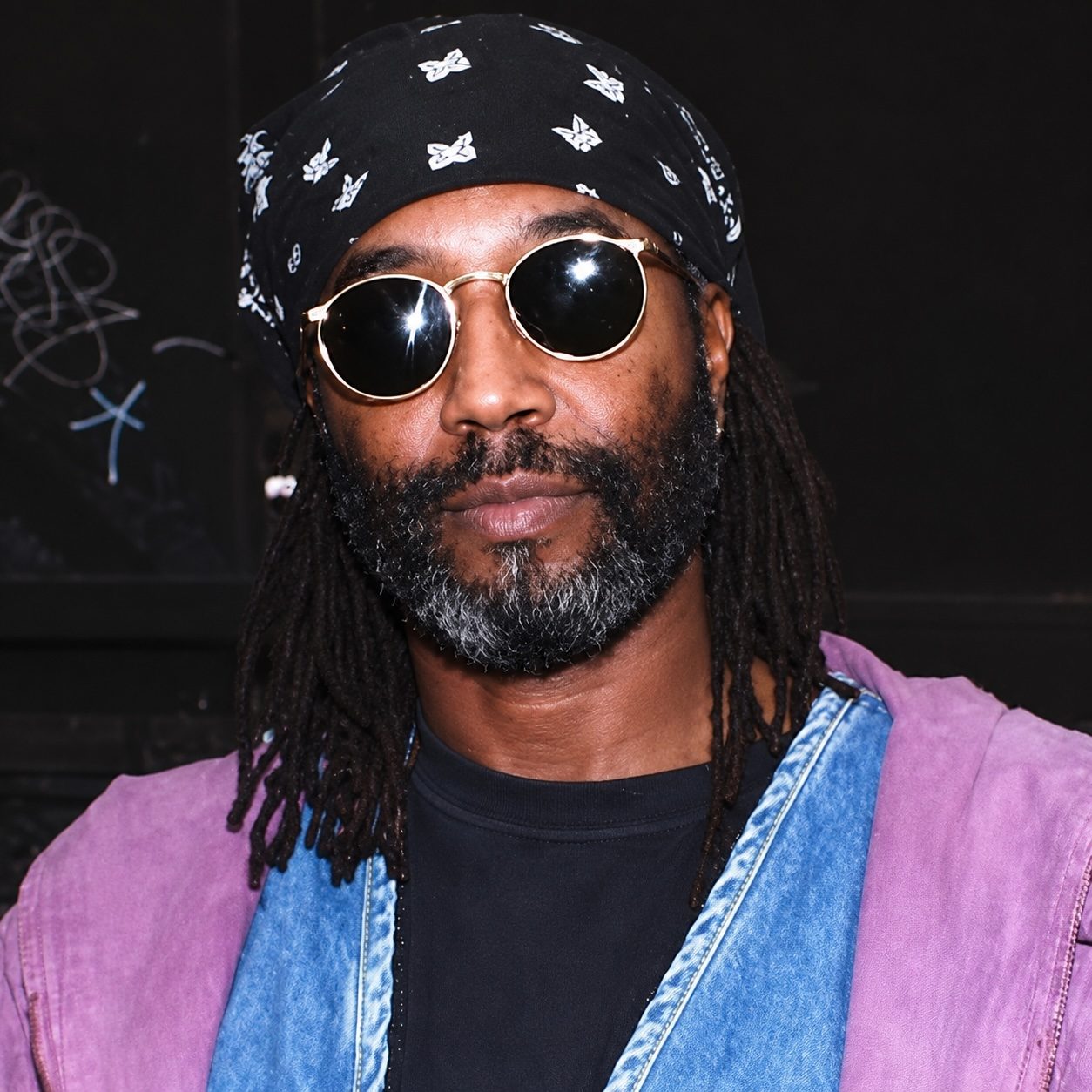 A man with long dreadlocks wearing sunglasses and a bandana, dressed in a colorful jacket, against a dark background.